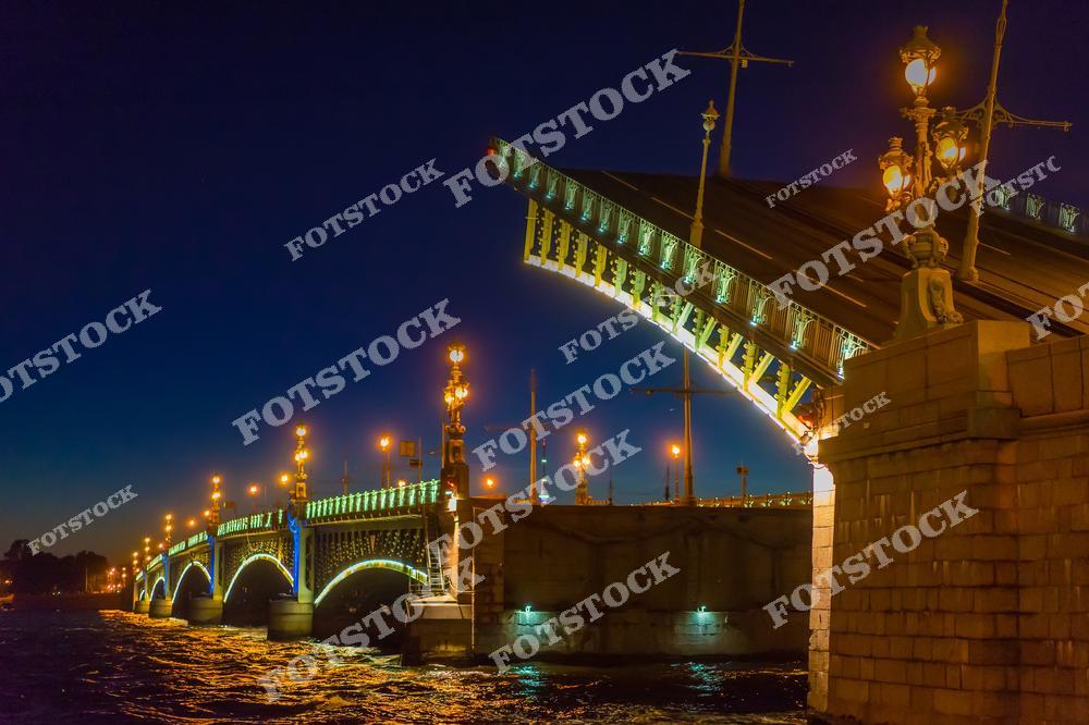 На фотографии изображён Троицкий мост в момент разведения, что является традиционным зрелищем для Санкт-Петербурга. Подсветка моста, вместе с изящными уличными фонарями, подчёркивает его архитектурные особенности, а свечение отражается в водах Невы. Этот мост, соединяющий центральные районы города, является важным архитектурным символом города на Неве.